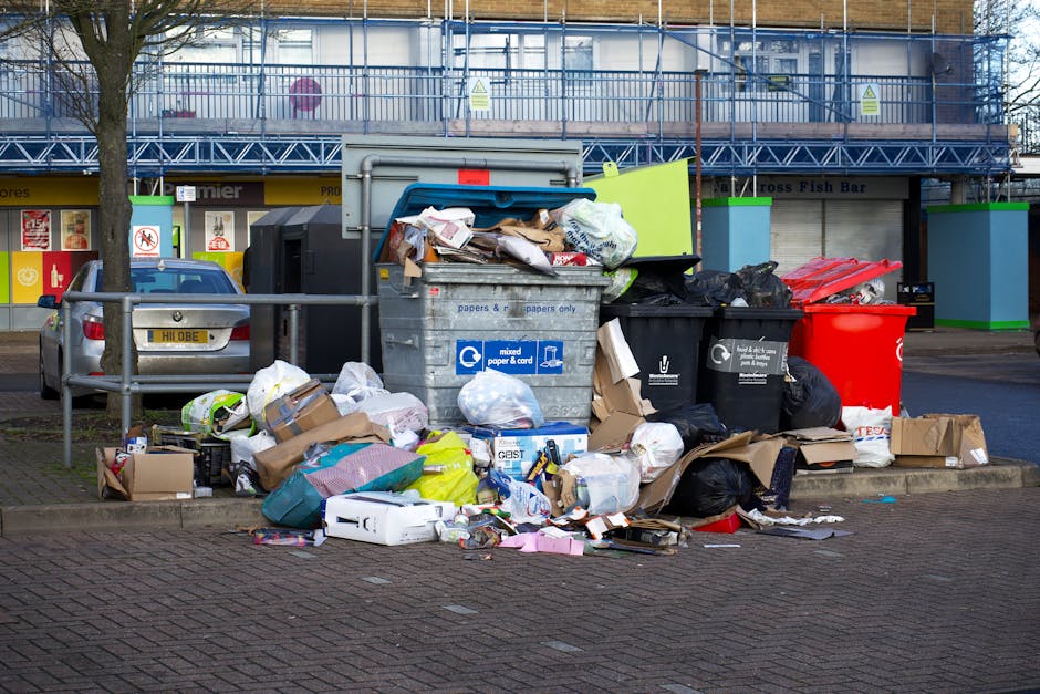 An outdoor scene showing a large collection of mixed waste and rubbish accumulated on a paved area near a street. The waste includes cardboard boxes, paper bags, black plastic garbage bags, and loose 