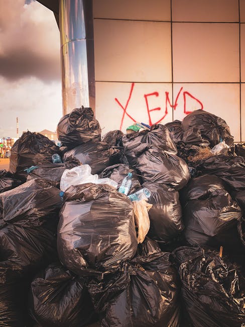 A large pile of black plastic rubbish bags, some tied tightly and others loosely open, stacked against a building with reflective metal panels. The bags contain mixed waste, with some transparent plas