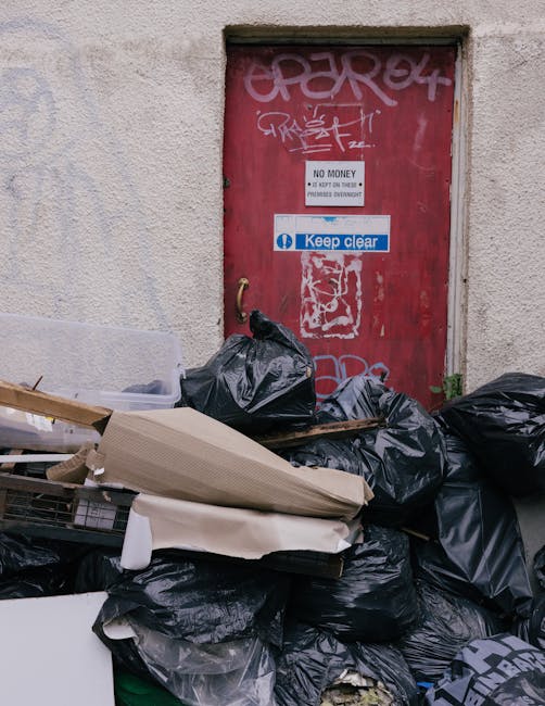 A collection of black plastic rubbish bags piled in front of a red metal door embedded in a concrete wall. The bags are tightly sealed and vary in size, with some appearing full and others partially o